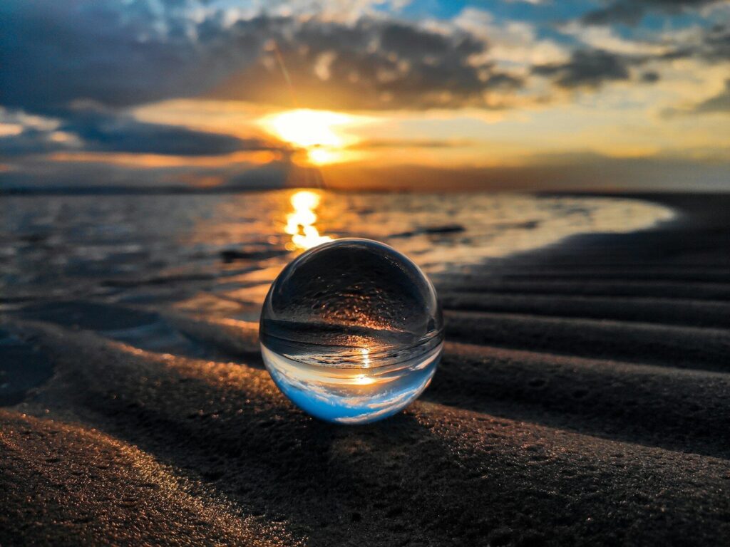 A glass sphere resting on sand reflecting a colorful sunset over a calm beach.